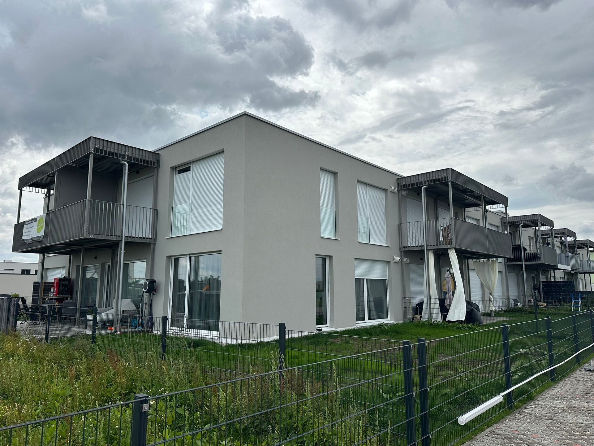 Moderne graue Stadthäuser mit Balkonen, bedeckter Himmel, hinter einem Maschendrahtzaun.