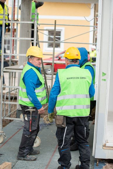 Eine Gruppe von Bauarbeitern in Westen und Schutzhelmen steht unter klarem Himmel vor einem Gerüst.