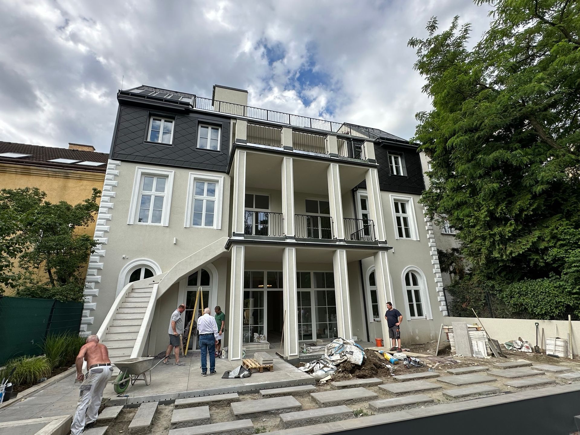 Baustelle eines zweistöckigen Gebäudes mit Arbeitern und Baumaterialien. Grau-weiße Fassade, blauer Himmel.