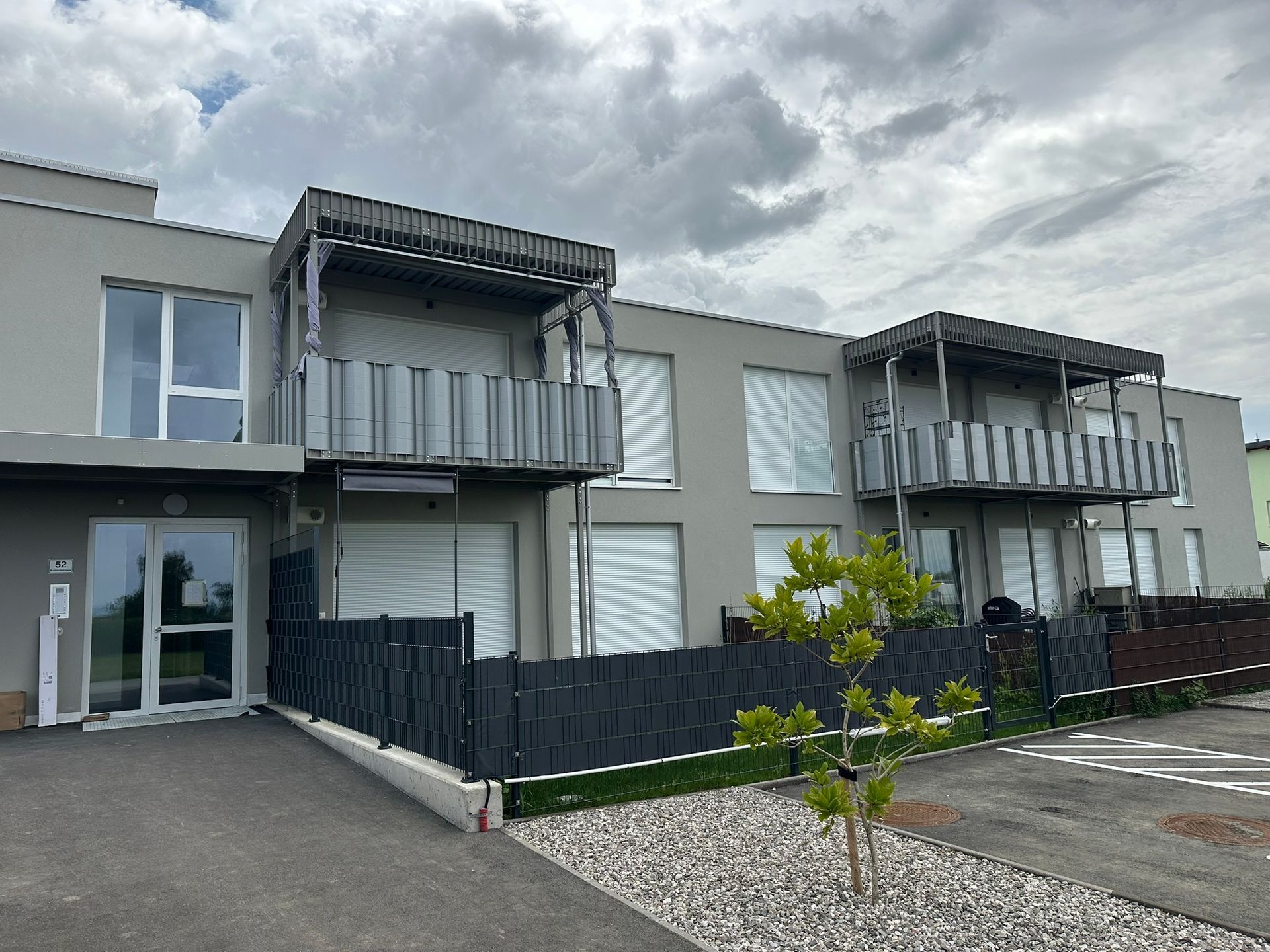 Graues Wohnhaus mit Balkonen, schwarzen Zäunen und einem kleinen Baum. Bewölkter Himmel.