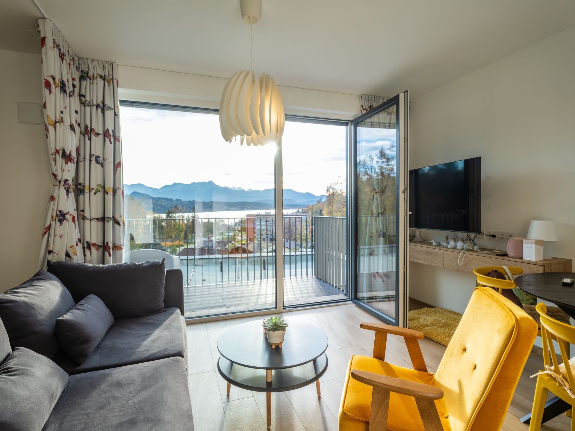 Wohnzimmer mit offenen Türen zu einem Balkon mit Blick auf einen See und Berge; graues Sofa, gelber Sessel und runder Tisch.