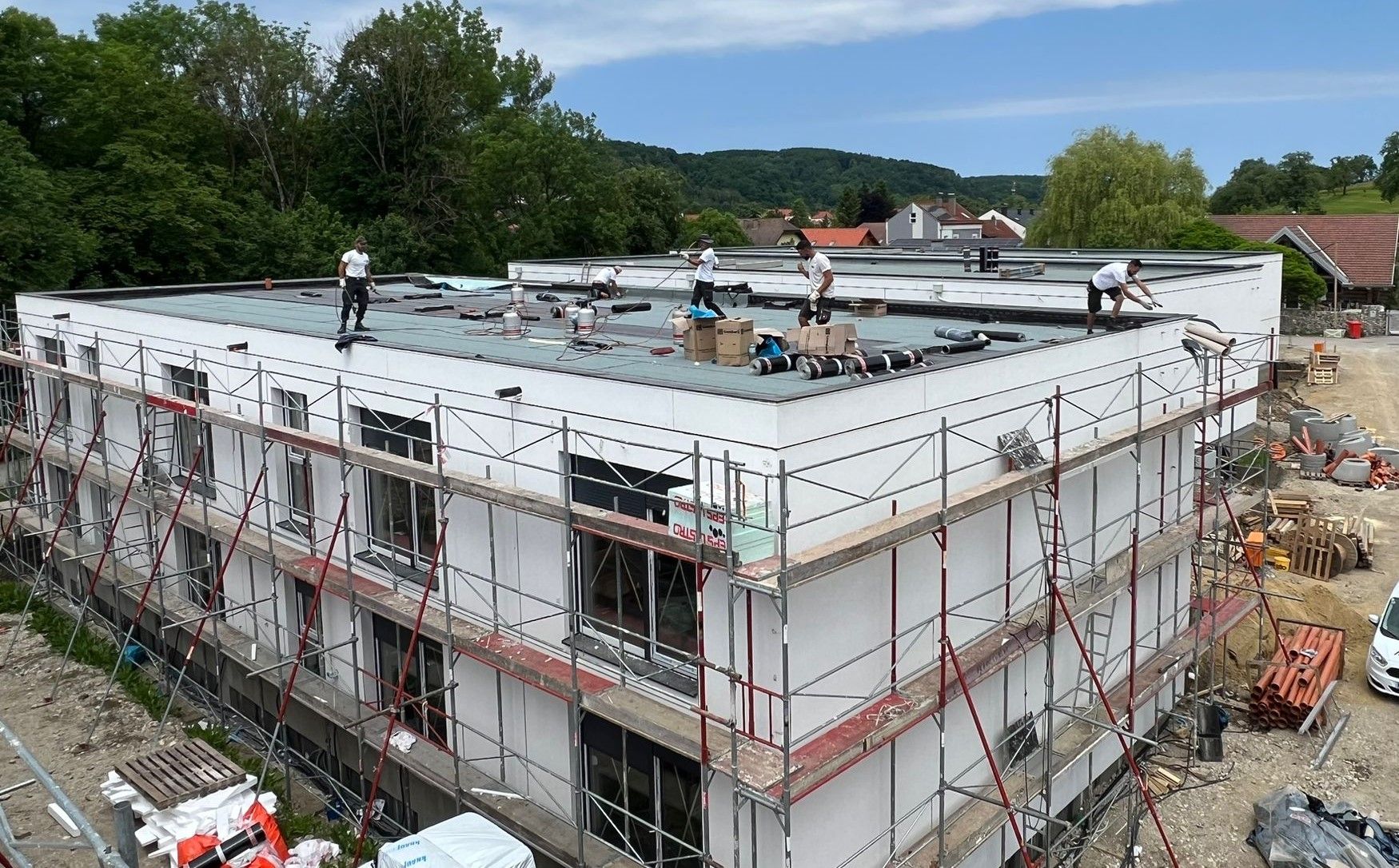 Bauarbeiter auf einem Flachdach errichten mit Gerüst ein mehrstöckiges Gebäude, umgeben von Bäumen und Baumaterialien.