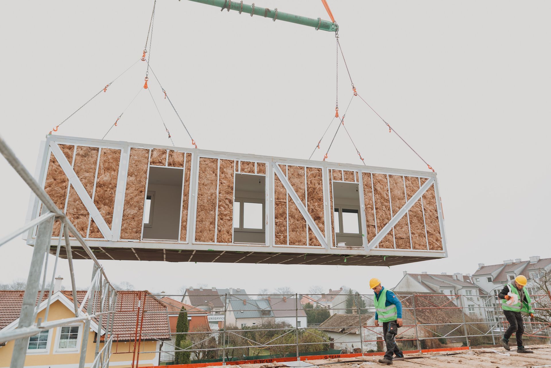 Eine modulare Baueinheit wird von einem Kran über eine Baustelle gehoben, Arbeiter tragen Sicherheitswesten.