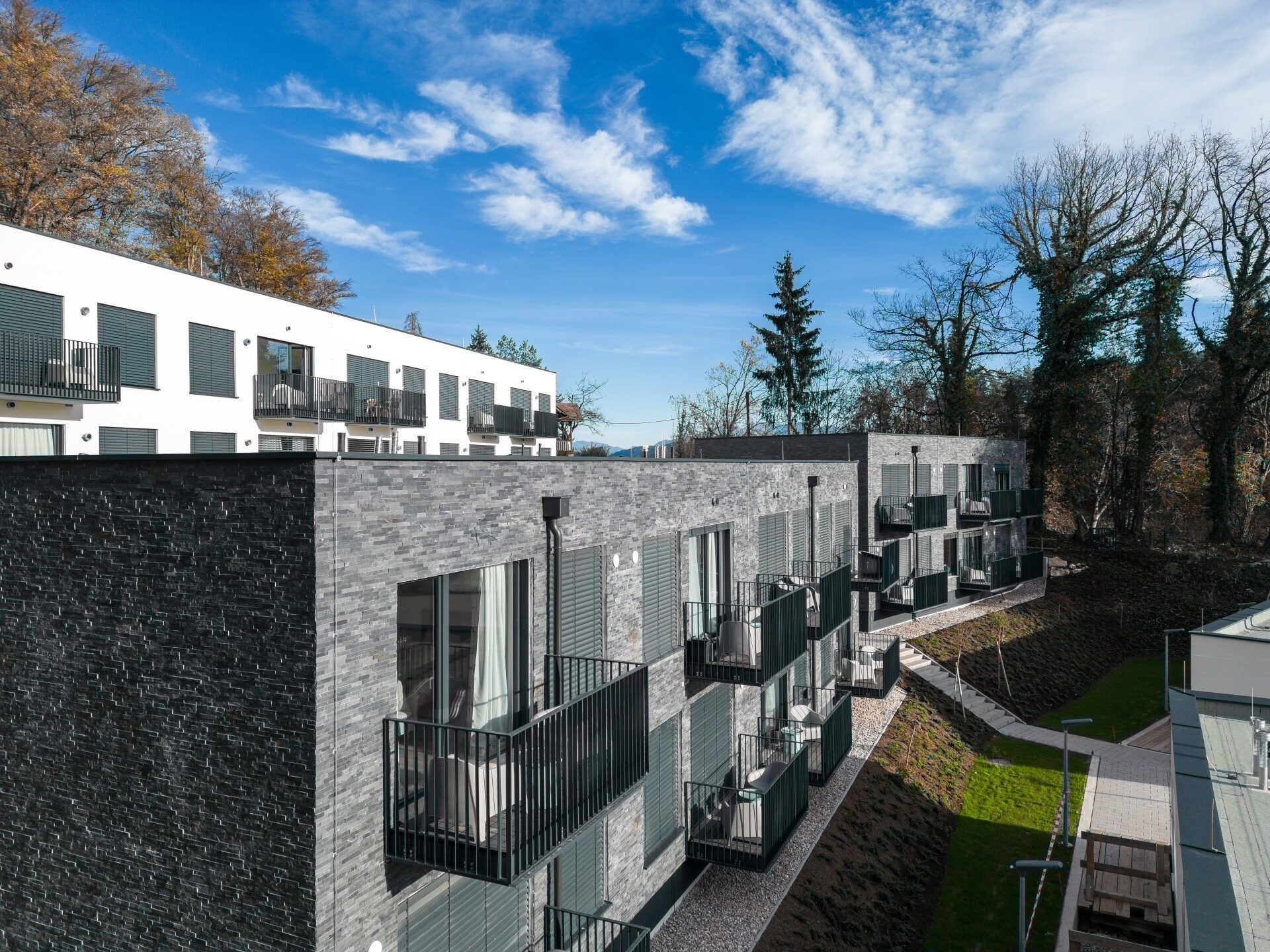 Moderne graue Backstein-Wohngebäude mit Balkonen, blauem Himmel und Bäumen im Hintergrund.