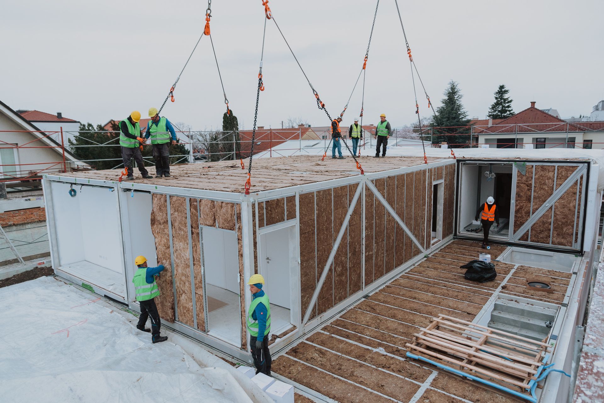 Bauarbeiter heben mit einem Kran ein Fertighaus auf ein schneebedecktes Dach. Gelbe Schutzhelme, blaue Westen.