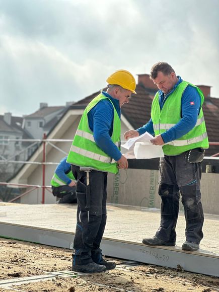 Eine Gruppe von Bauarbeitern in Westen und Schutzhelmen steht unter klarem Himmel vor einem Gerüst.