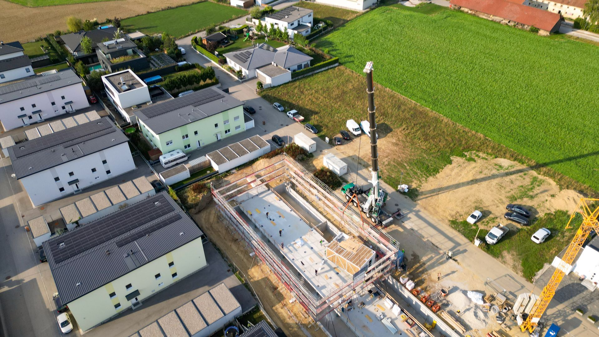 Luftaufnahme: Baustelle mit Kran, in der Nähe von Wohnhäusern und einem Feld.