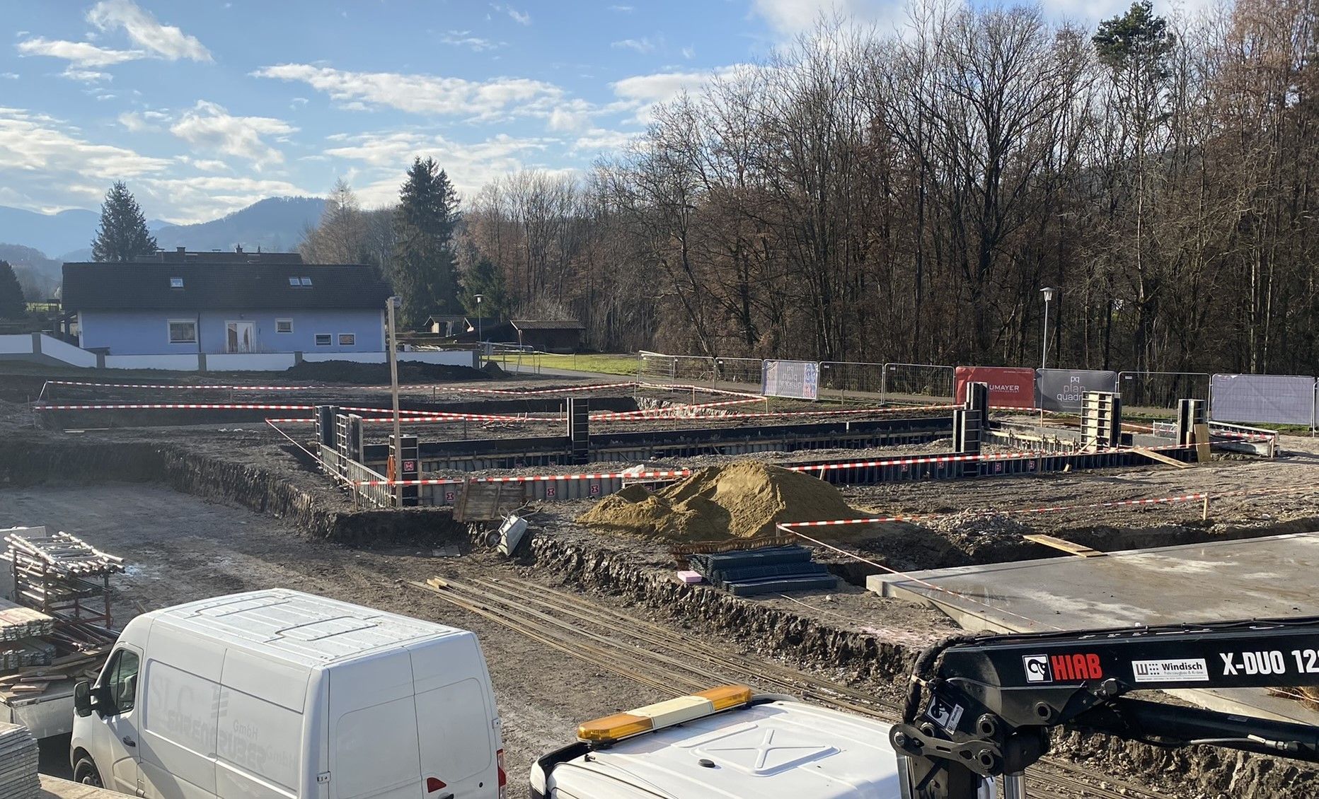 Baustelle mit ausgegrabenen Fundamenten, weißem Lieferwagen und bewaldetem Hintergrund.