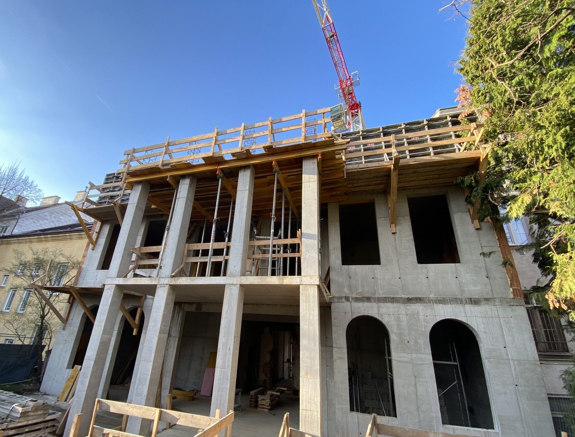 Im Bau befindliches Gebäude mit Sichtbetonrahmen, Holzgerüst und einem Kran vor blauem Himmel.