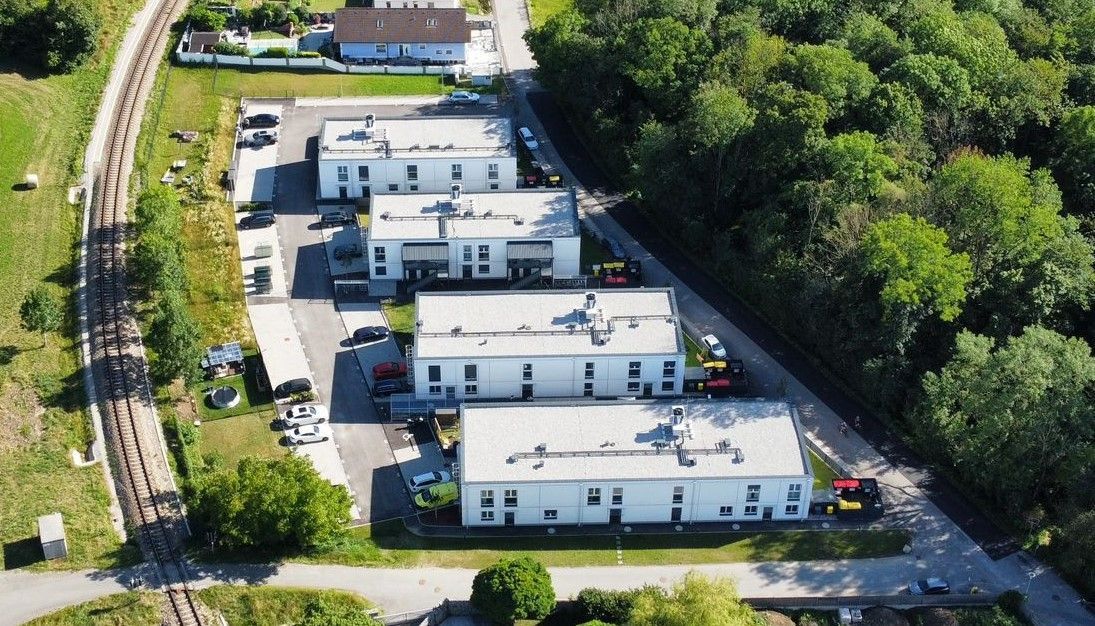 Eine Luftaufnahme moderner weißer Gebäude mit grauen Dächern neben einer Bahnstrecke und einem Wald.