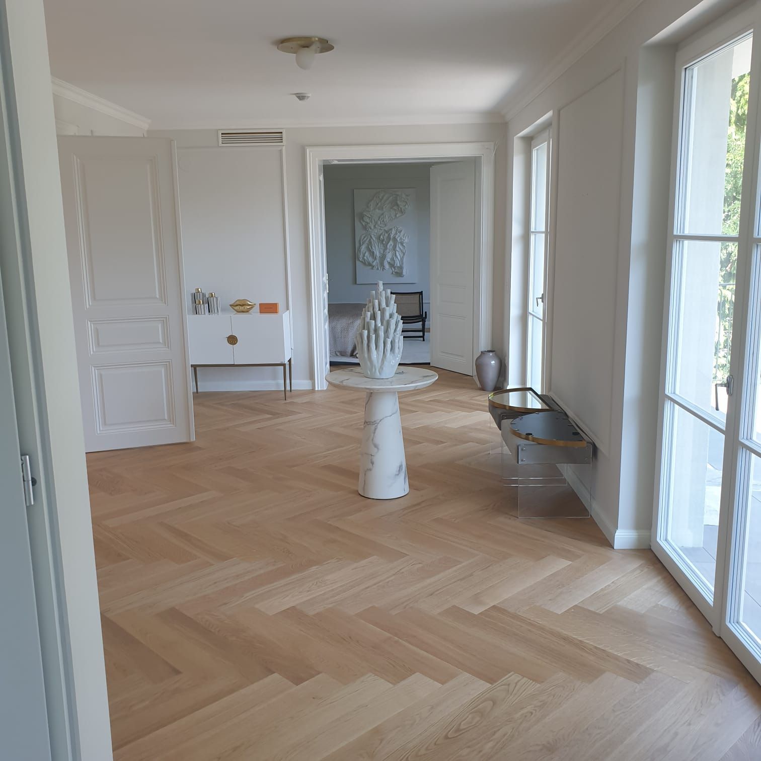Heller, lichtdurchfluteter Innenflur mit Fischgrätholzböden, weißen Wänden und einem dekorativen Säulentisch.