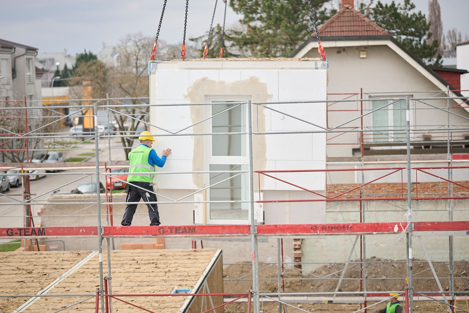 Bauarbeiter auf Gerüst, der ein vorgefertigtes Wandelement mit einem Fenster führt, das von einem Kran angehoben wird.