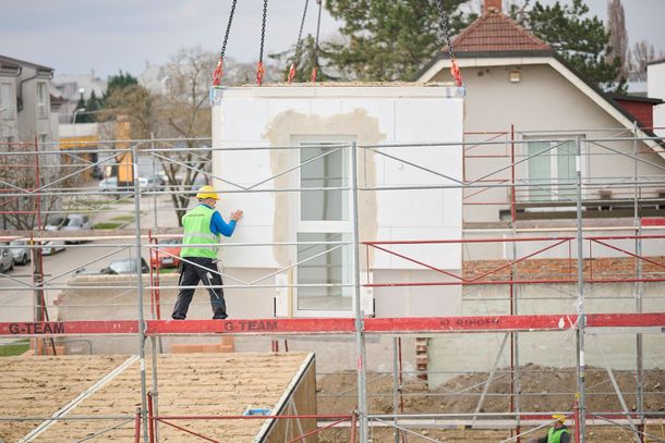 Bauarbeiter auf Gerüst, der ein vorgefertigtes Wandelement mit einem Fenster führt, das von einem Kran angehoben wird.