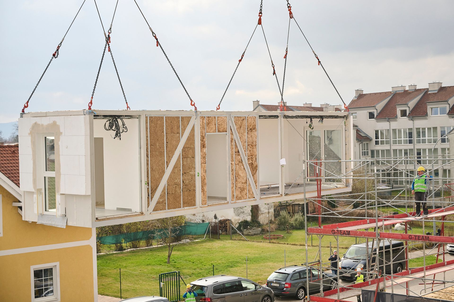 Ein vorgefertigtes Hausmodul wird mit einem Kran auf ein im Bau befindliches Gebäude gehoben; im Freien.
