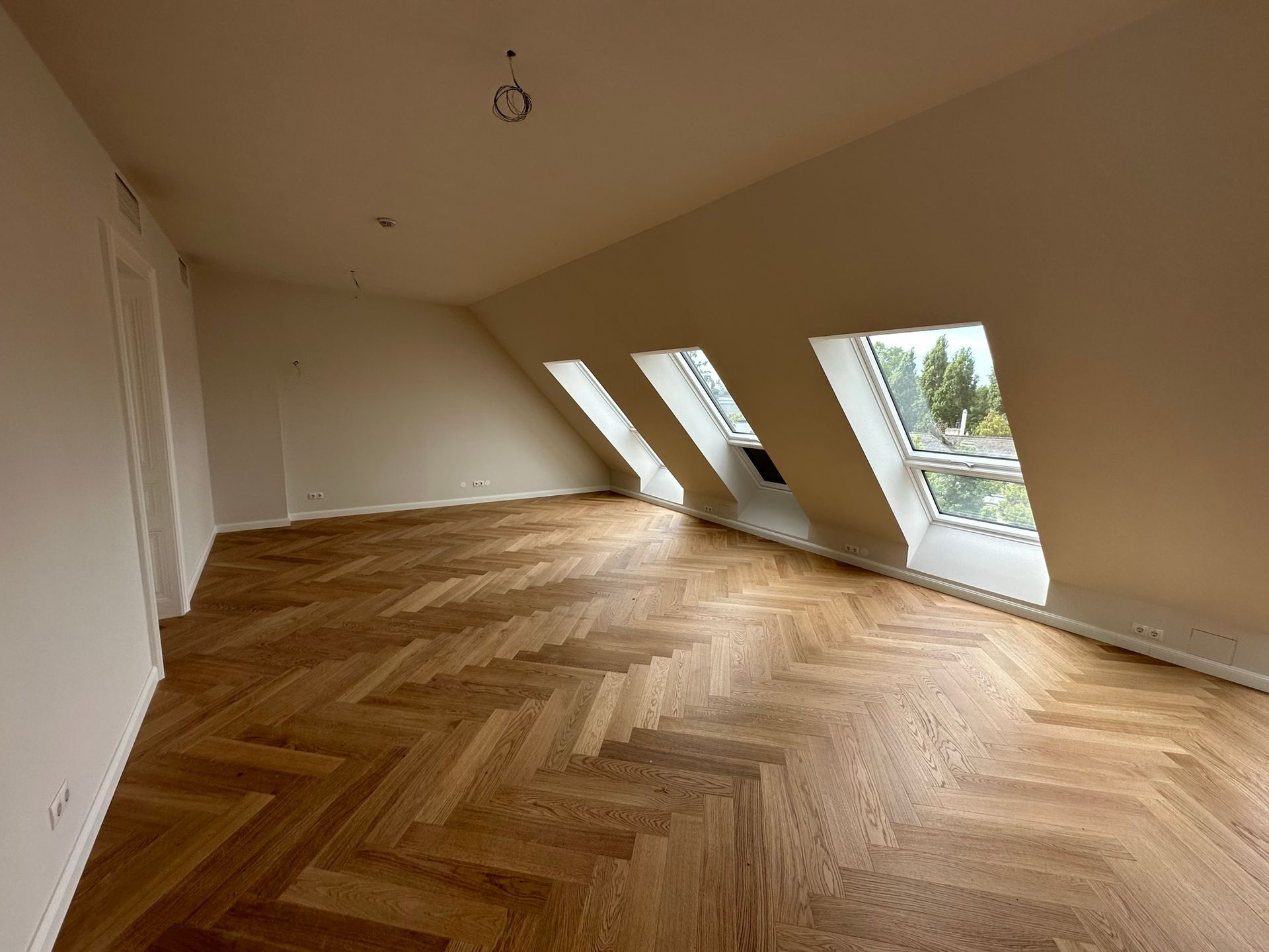 Leerer Raum mit schräger Decke, drei Dachfenstern und Holzboden im Fischgrätenmuster.