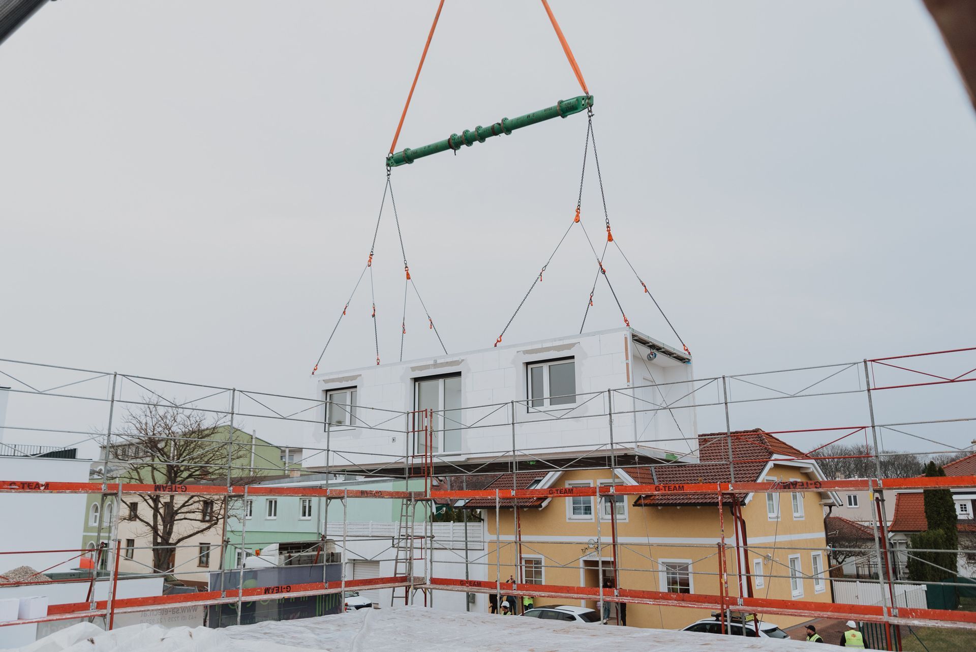 Ein modulares Gebäude wird von einem Kran auf ein Dach gehoben, umgeben von Gerüsten und Wohngebäuden.