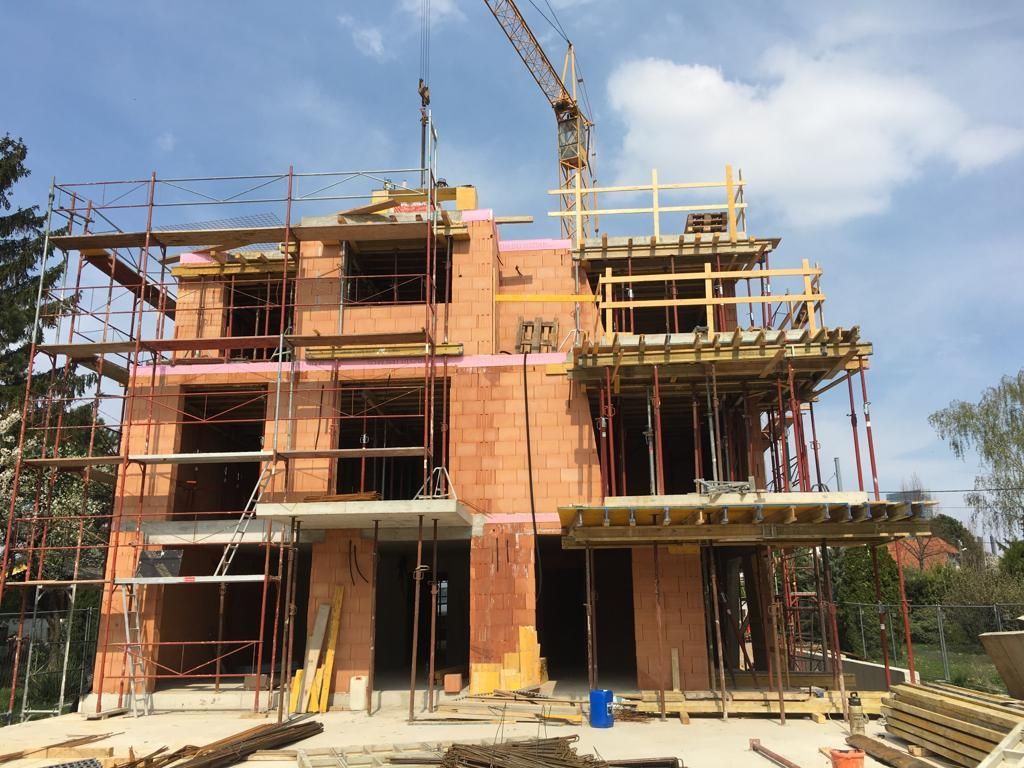 Bau eines zweistöckigen Backsteinhauses mit Kran, Gerüst und Holzschalungen vor blauem Himmel.