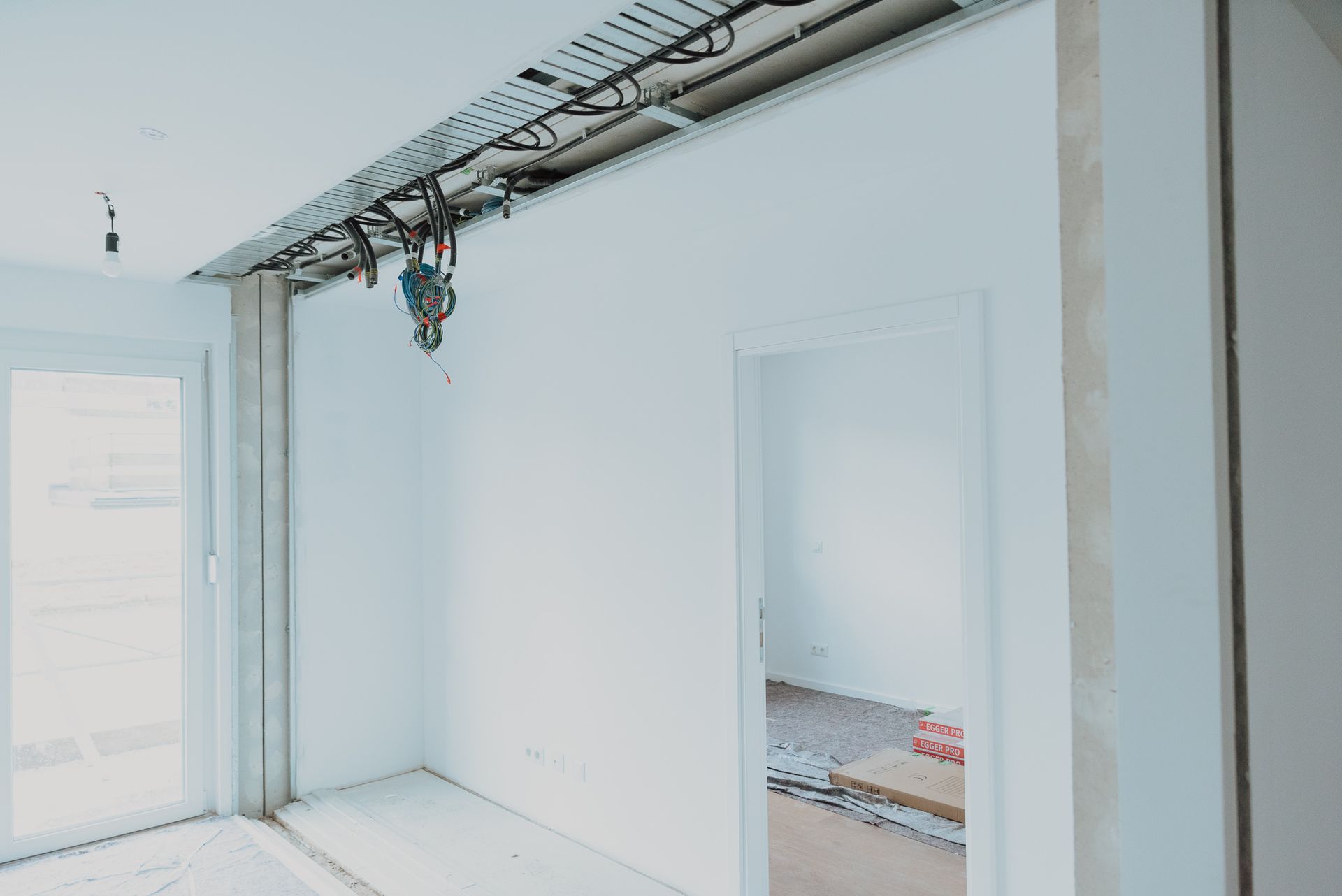Innenansicht eines im Bau befindlichen Raums mit freiliegenden Kabeln, einer Tür und einem Fenster.