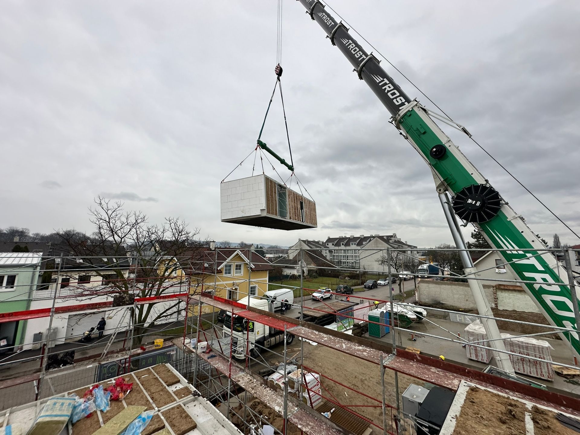 Ein Kran hebt ein rechteckiges modulares Gebäudeelement über eine Baustelle, auf der andere Gebäude sichtbar sind.