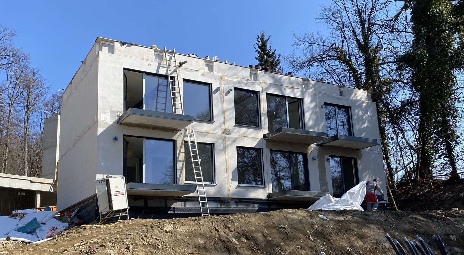 Zweistöckiges Gebäude im Bau mit reflektierenden Fenstern vor einer sonnigen Hügelkulisse.