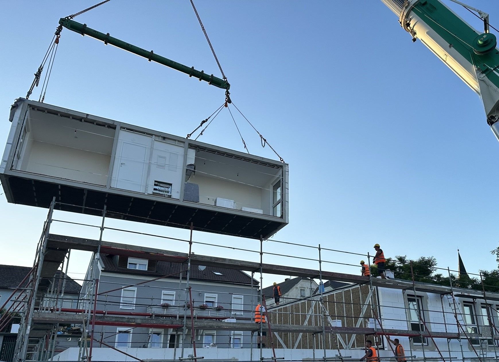 Ein Kran hebt eine modulare Baueinheit auf eine Baustelle; Arbeiter in orangefarbenen Westen sind zu sehen.