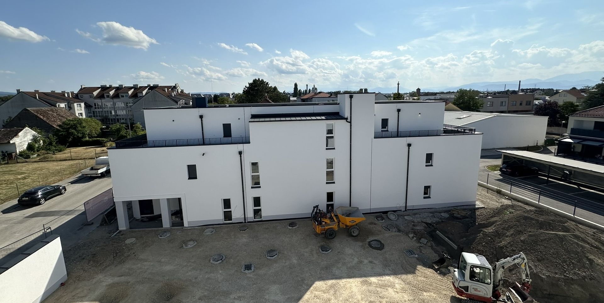 Ein weißes, modernes Gebäude im Bau mit Erdbewegungsmaschinen. Häuser und ein blauer Himmel mit Wolken im Hintergrund.