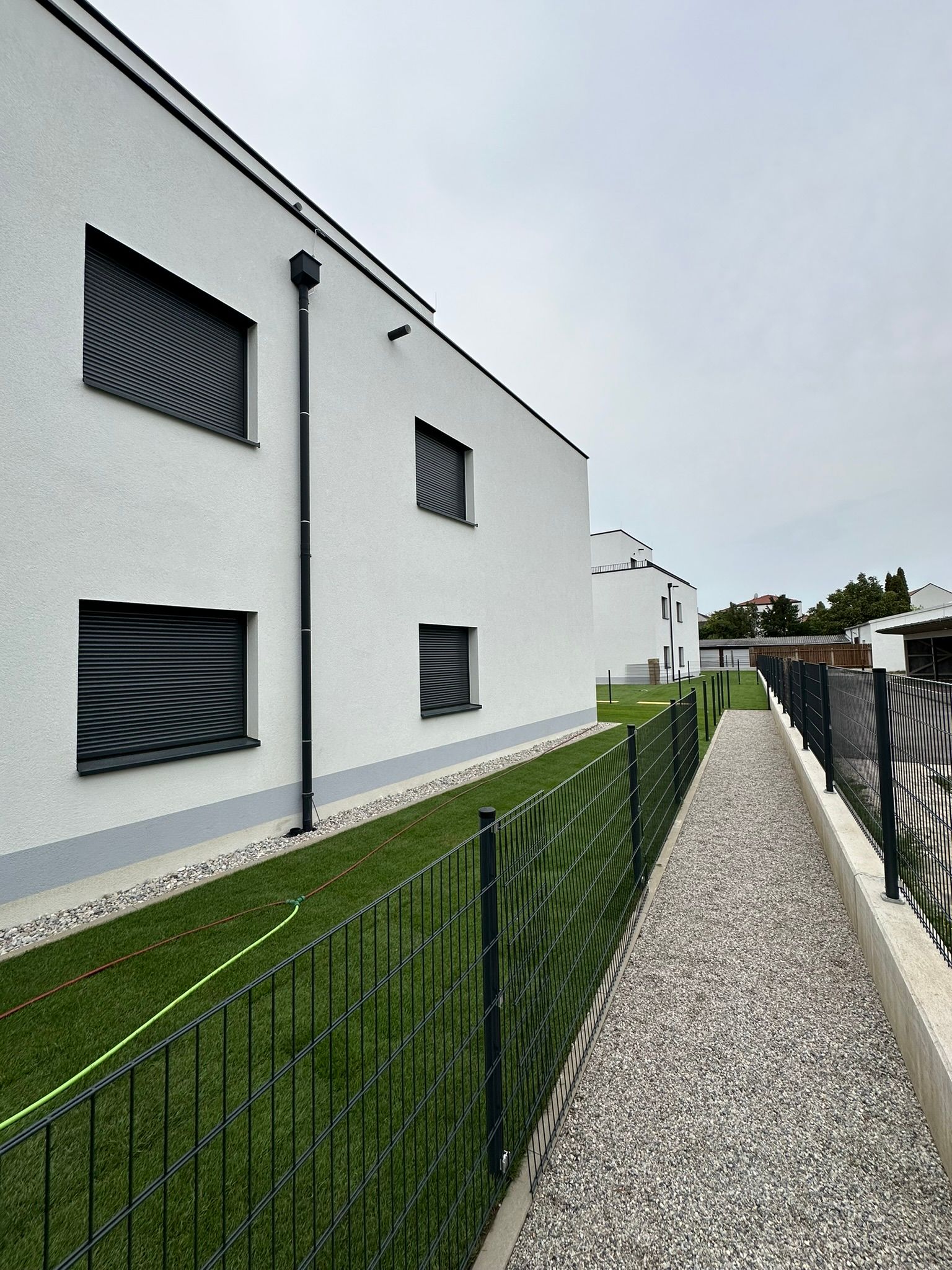 Weiße, moderne Gebäude mit dunklen Jalousien und einem schwarzen Zaun, der an einen Kiesweg und Gras grenzt.