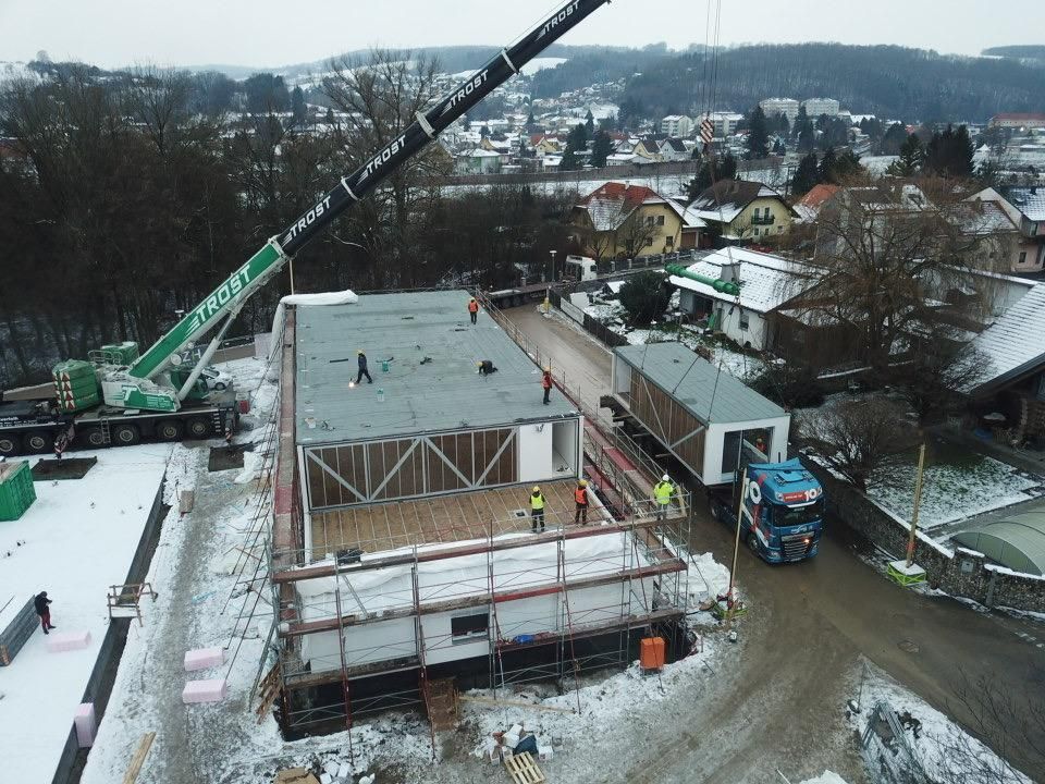 Ein Kran hebt ein vorgefertigtes Gebäudemodul auf einer Baustelle in einer verschneiten Umgebung an.