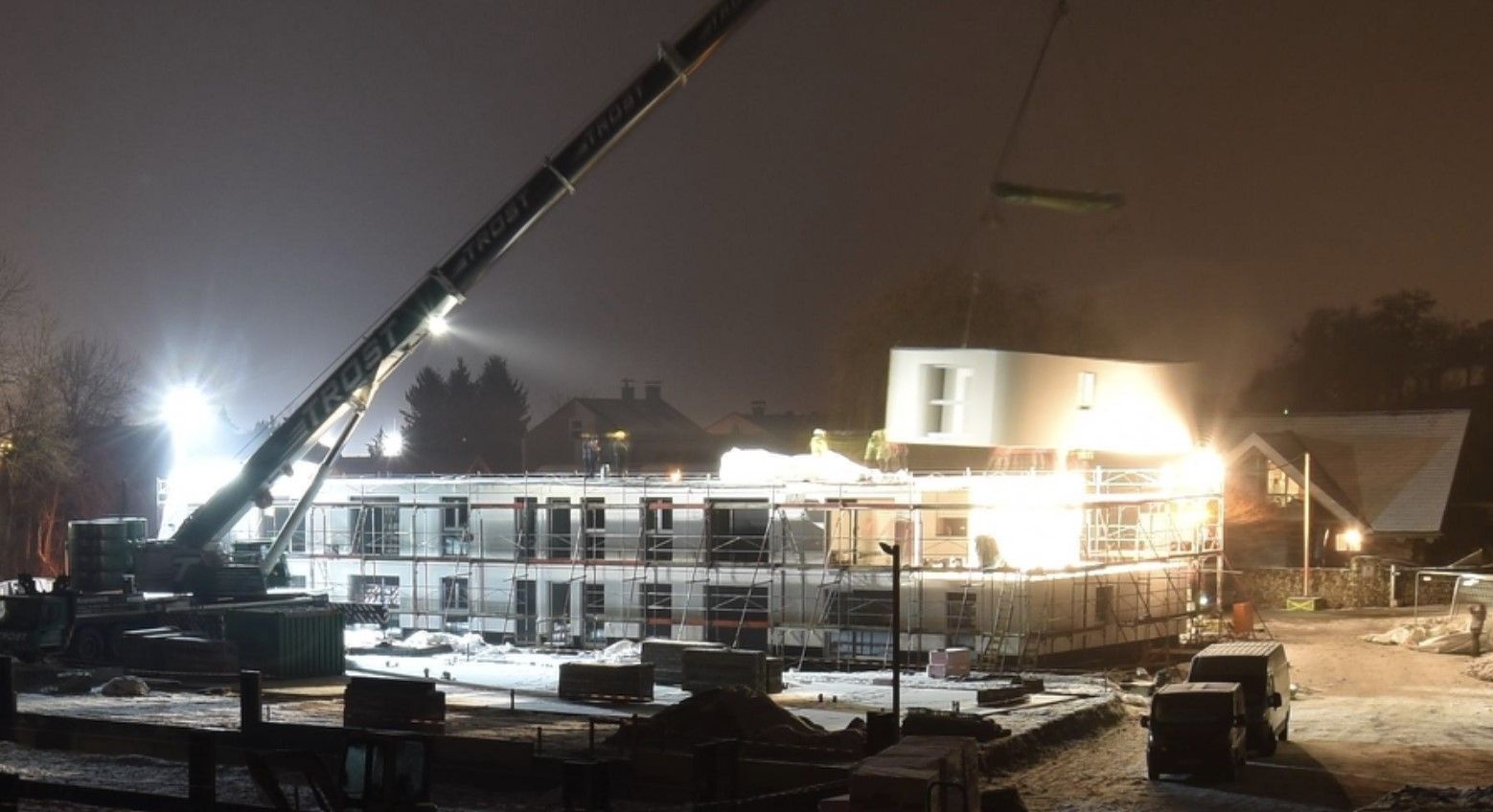 Baustelle bei Nacht; Kran hebt eine modulare Baueinheit auf eine teilweise errichtete Struktur.