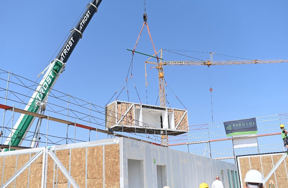 Baustelle mit Kran, der modulare Baueinheit anhebt. Blauer Himmel, Arbeiter und andere Bauelemente sichtbar.