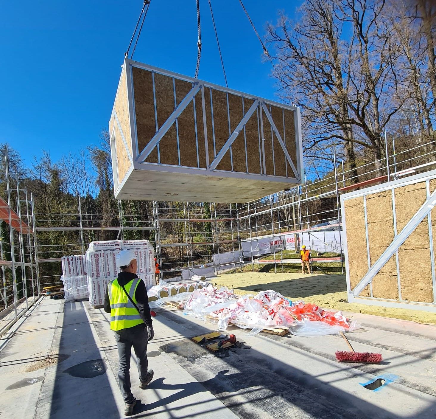 Eine modulare Baueinheit wird auf einer Baustelle an ihren Platz abgesenkt; ein Arbeiter schaut zu.