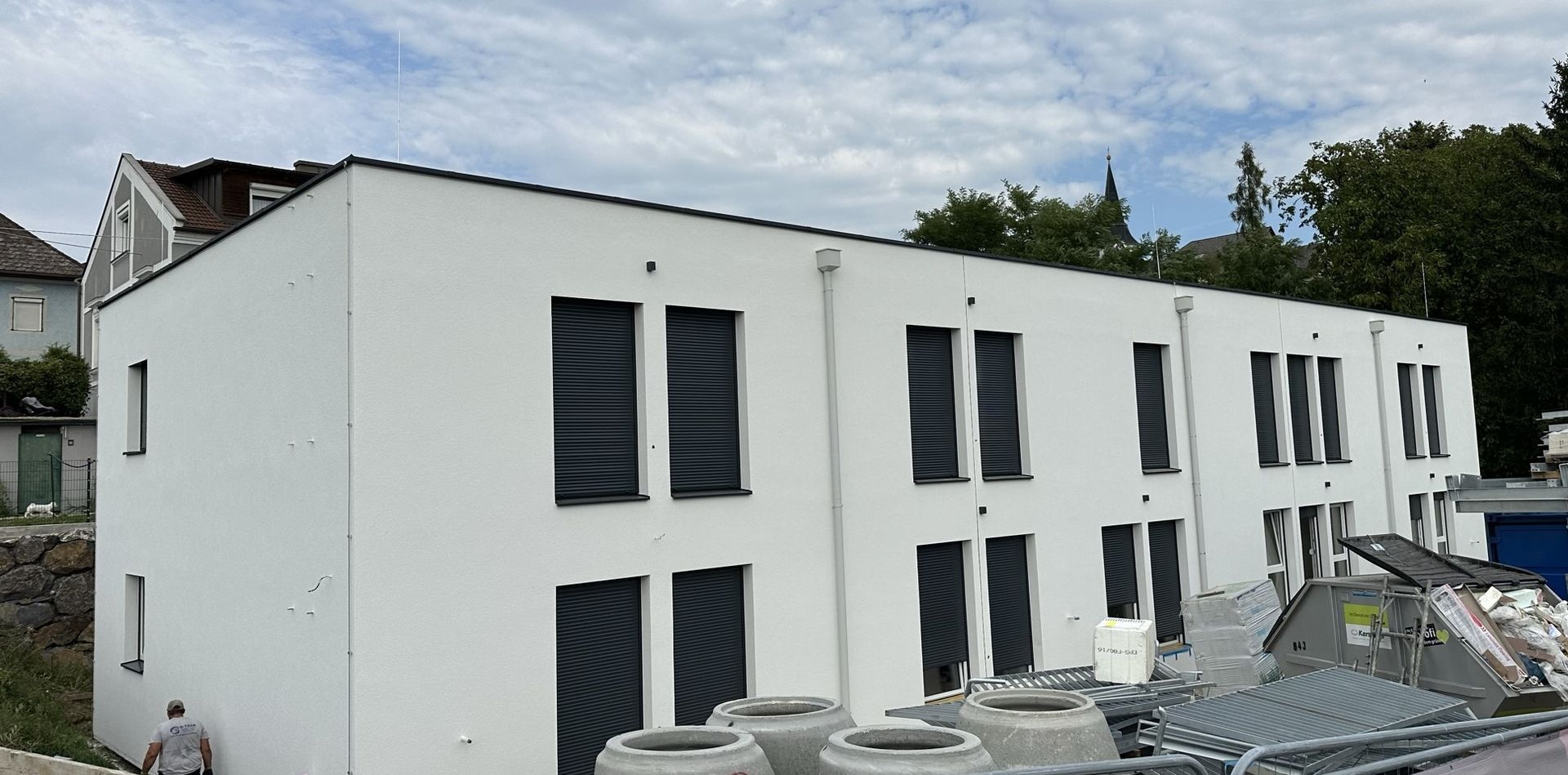 Weißes Mehrfamilienhaus mit schwarzen Fensterläden unter bewölktem Himmel.