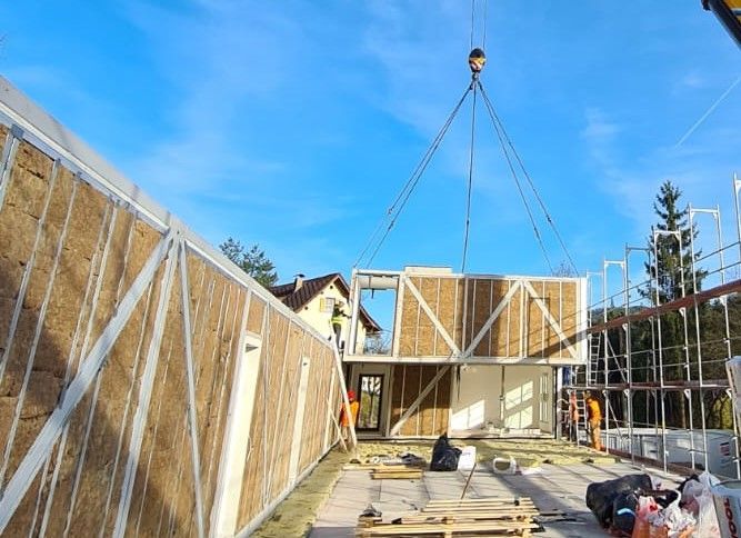 Baustelle: Kran hebt einen modularen Gebäudeabschnitt an. Arbeiter in orangefarbenen Westen, blauer Himmel.