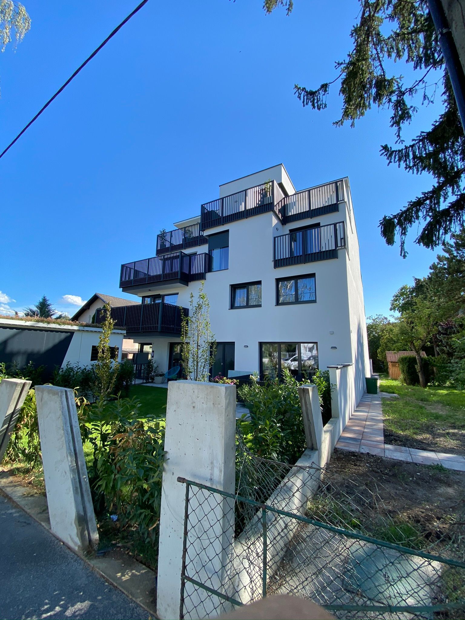 Weißes Mehrfamilienhaus mit Balkonen, grünem Rasen und einem Zaun an einem sonnigen Tag.
