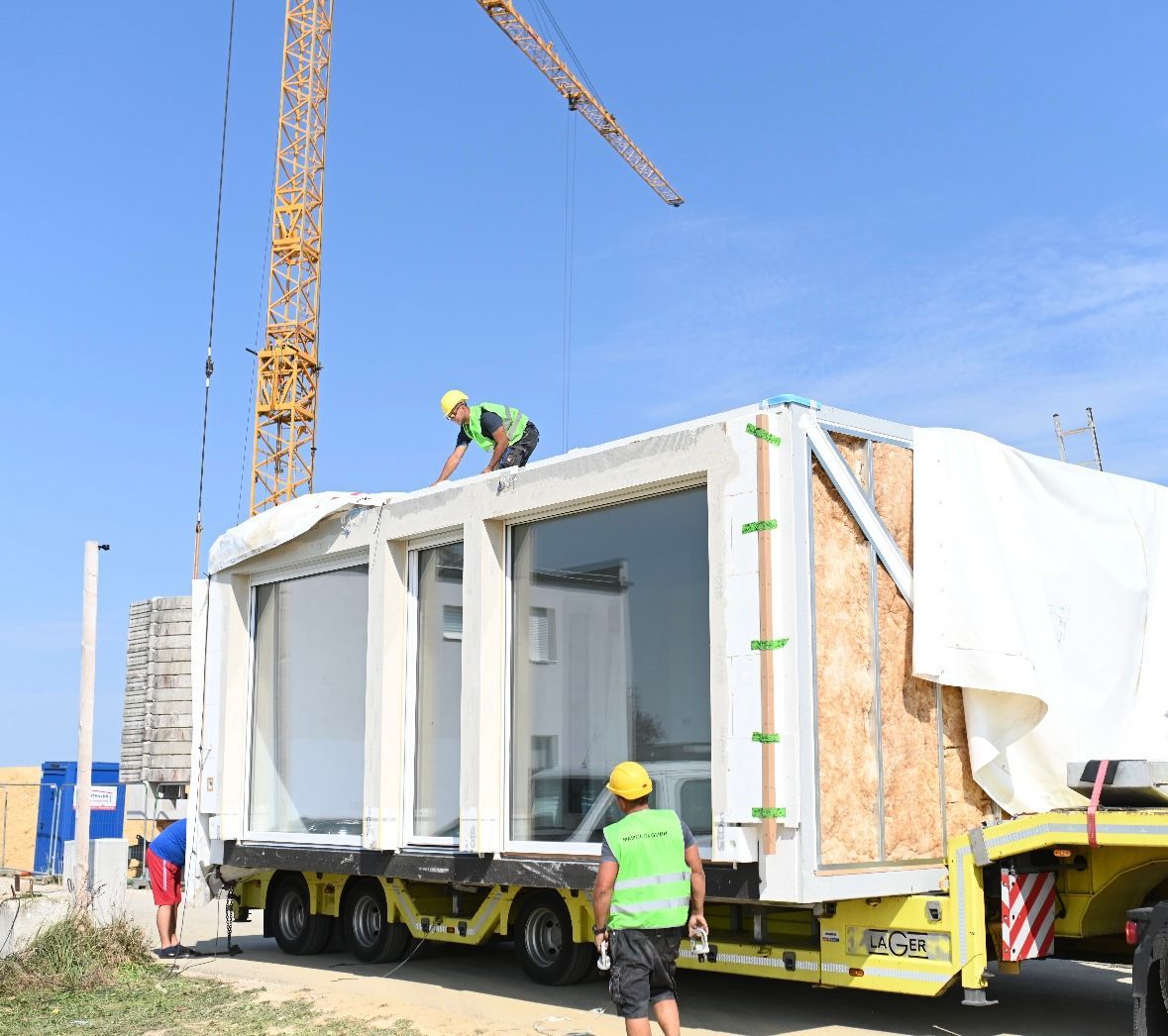 Arbeiter auf einem Pritschenwagen bewegen mit einem Kran ein modulares Gebäude mit großen Fenstern auf einer Baustelle.