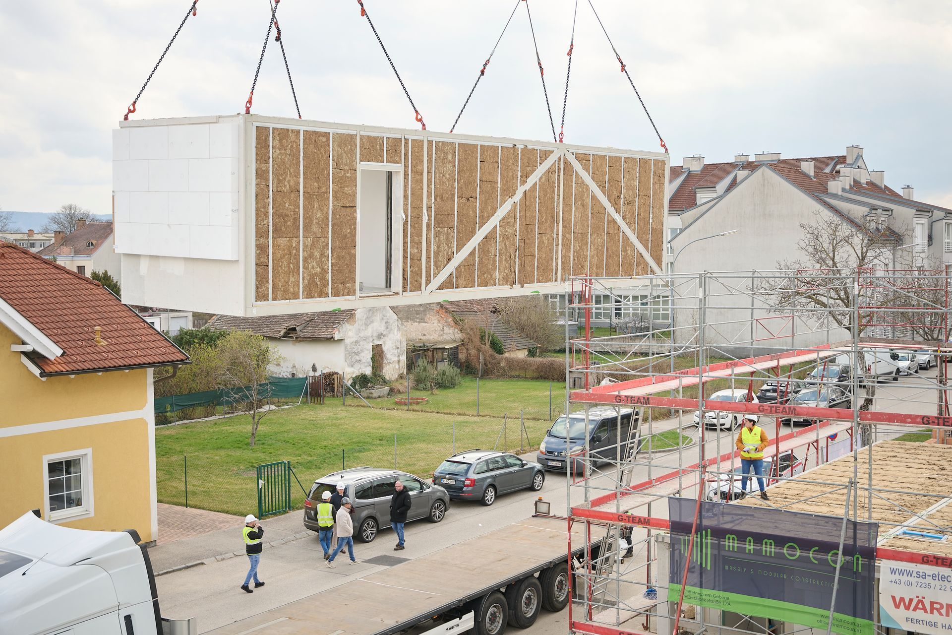 Ein Kran hebt eine modulare Gebäudeeinheit über eine Straße mit Fahrzeugen und Bauarbeitern.