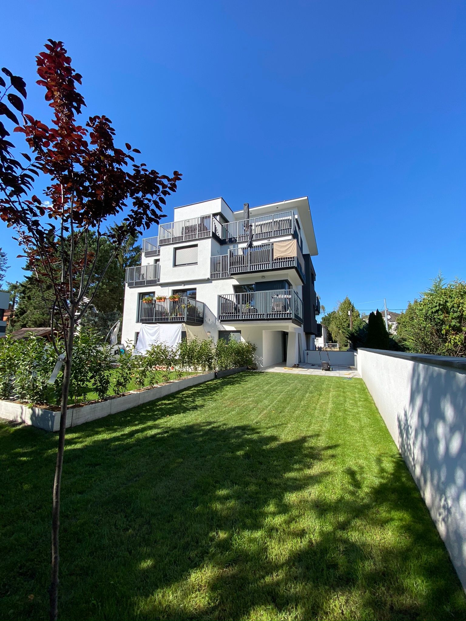Modernes weißes Wohnhaus mit Balkonen, grünem Rasen und strahlend blauem Himmel.