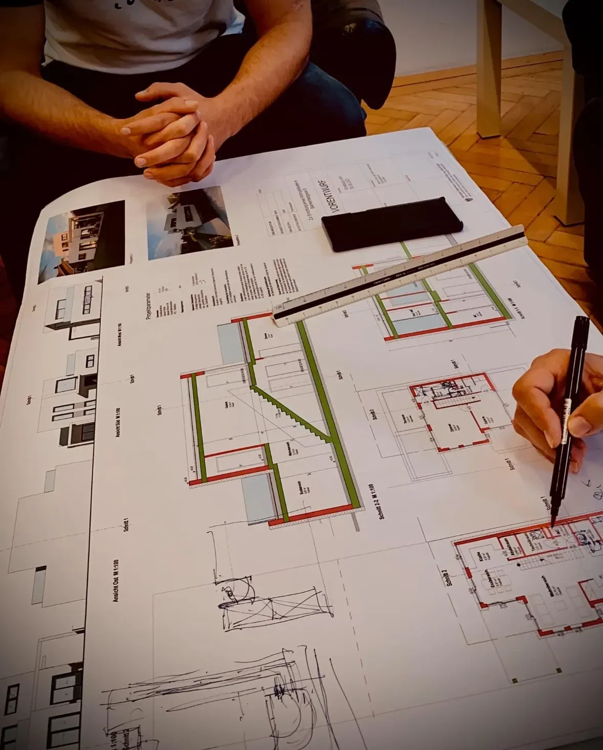 Zwei Personen an einem Tisch prüfen Baupläne, eine Person zeichnet mit einem Stift.
