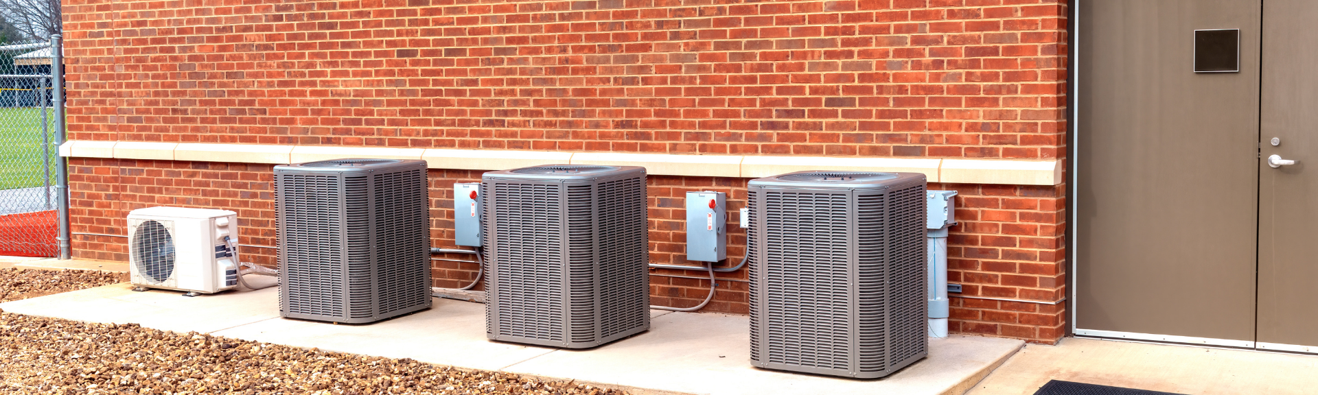 Three industrial HVAC air conditioning units sit on a concrete pad against a brick wall next to a door.