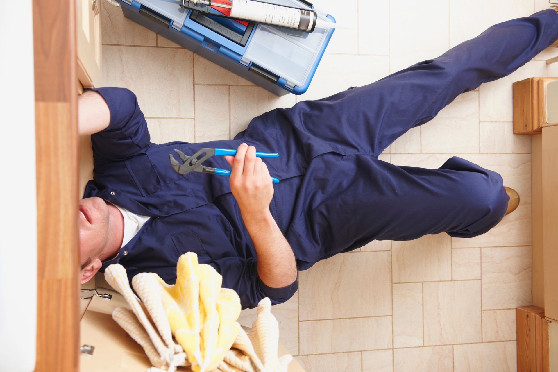 A plumber in blue overalls fixing a cabinet while lying on the floor, tools nearby