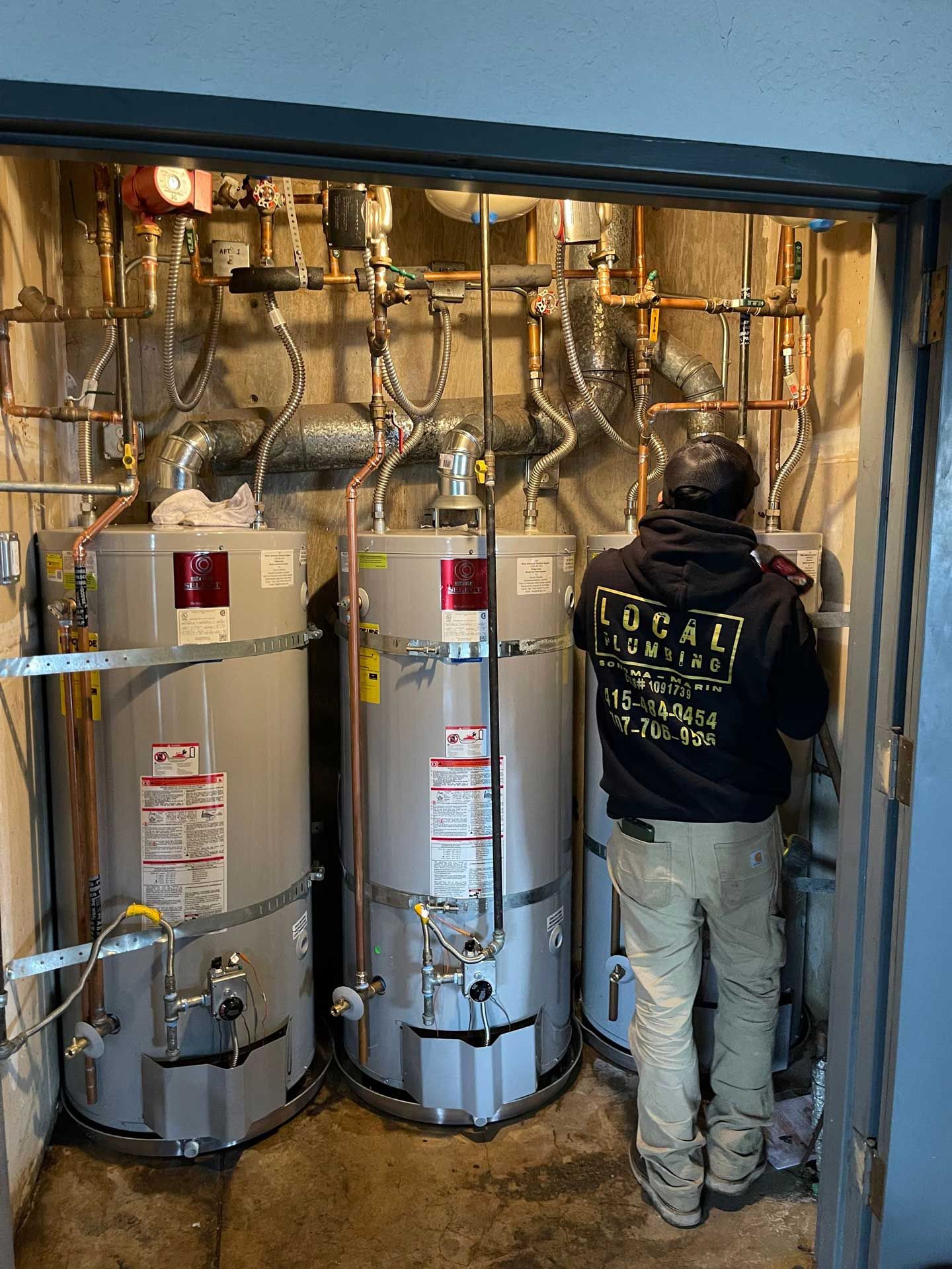Plumber working on three water heaters in a utility room. Copper pipes and wiring are visible.