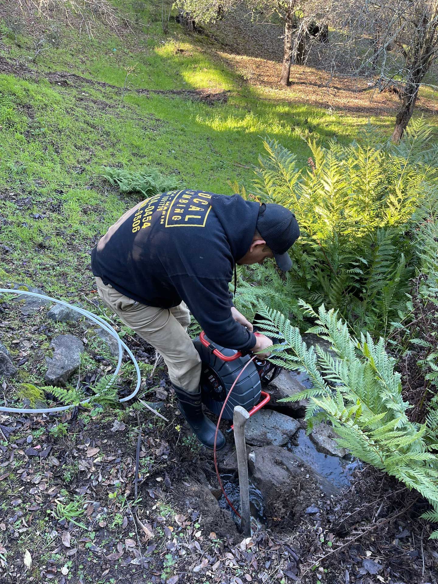 Person in black hoodie working on machinery next to small stream in forest setting.