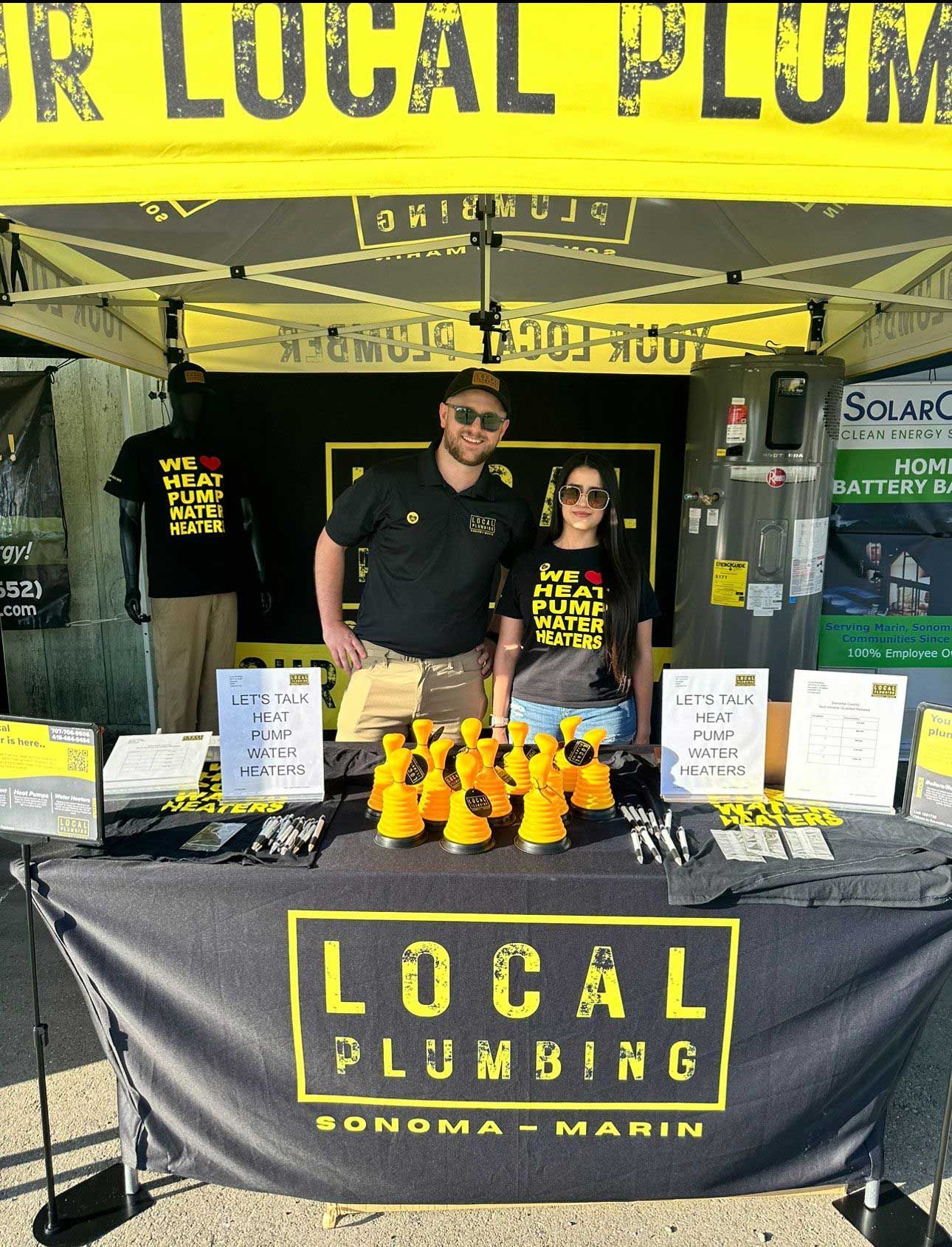 A Local Plumbing booth at an outdoor event. Two people stand behind a table with merchandise and a banner.