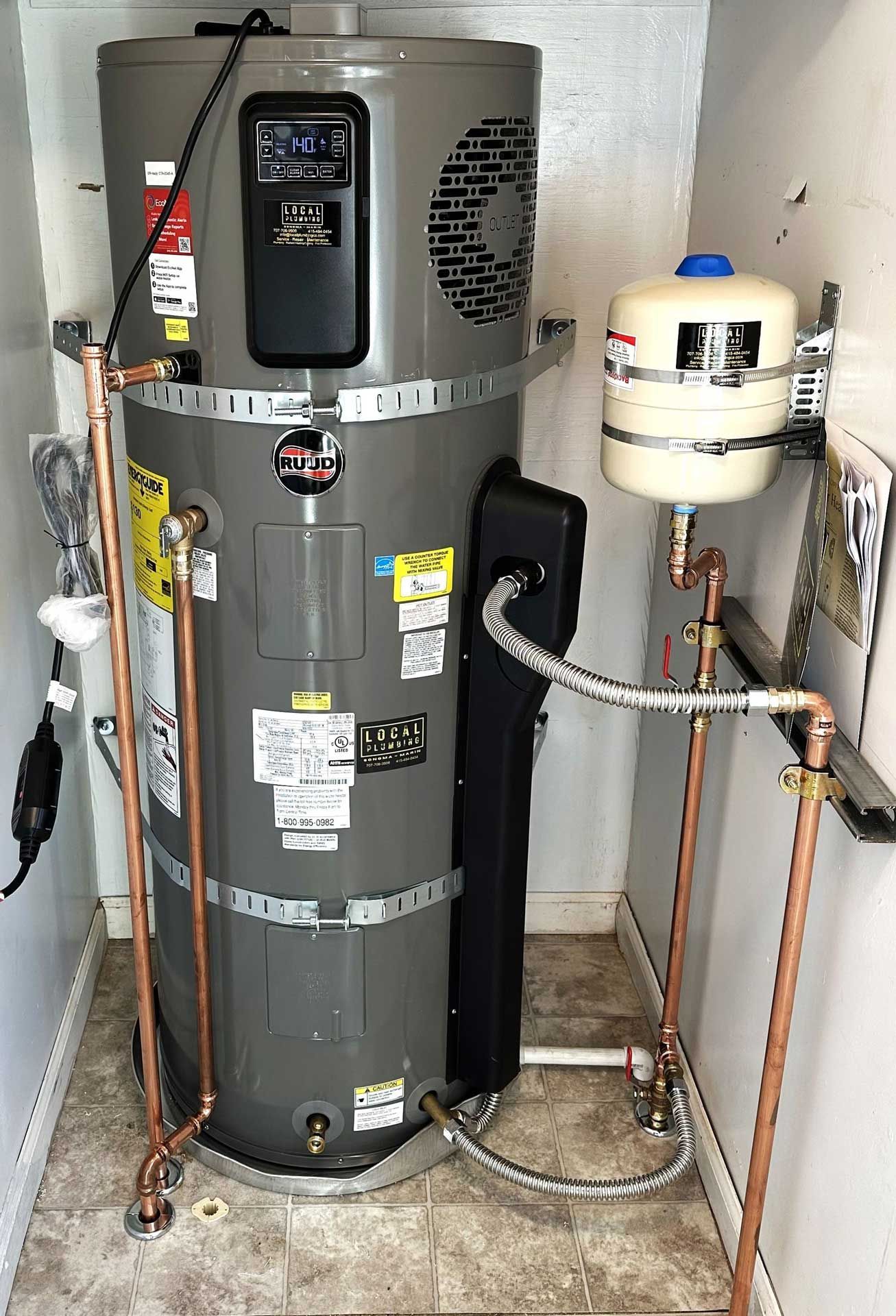 A tall, gray water heater with copper pipes in a utility closet, next to an expansion tank.