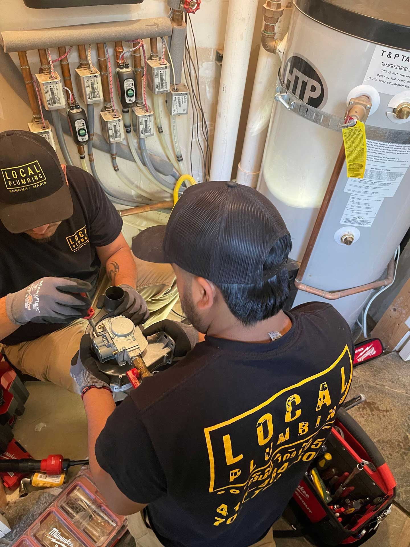 Two plumbers working on pipes near a water heater in a utility room. One uses a grinder.
