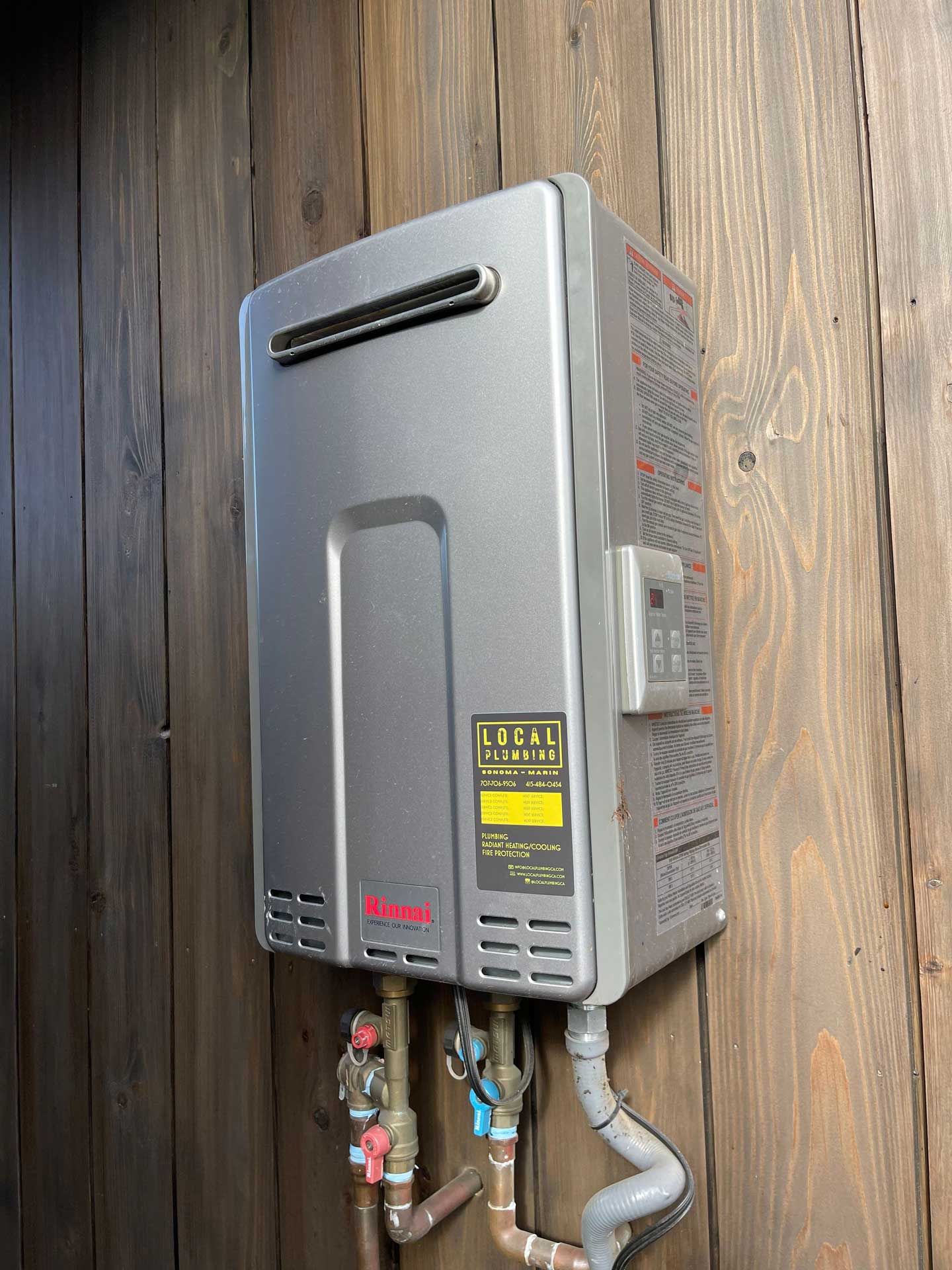 Silver tankless water heater mounted on wood paneling, with pipes and control panel.