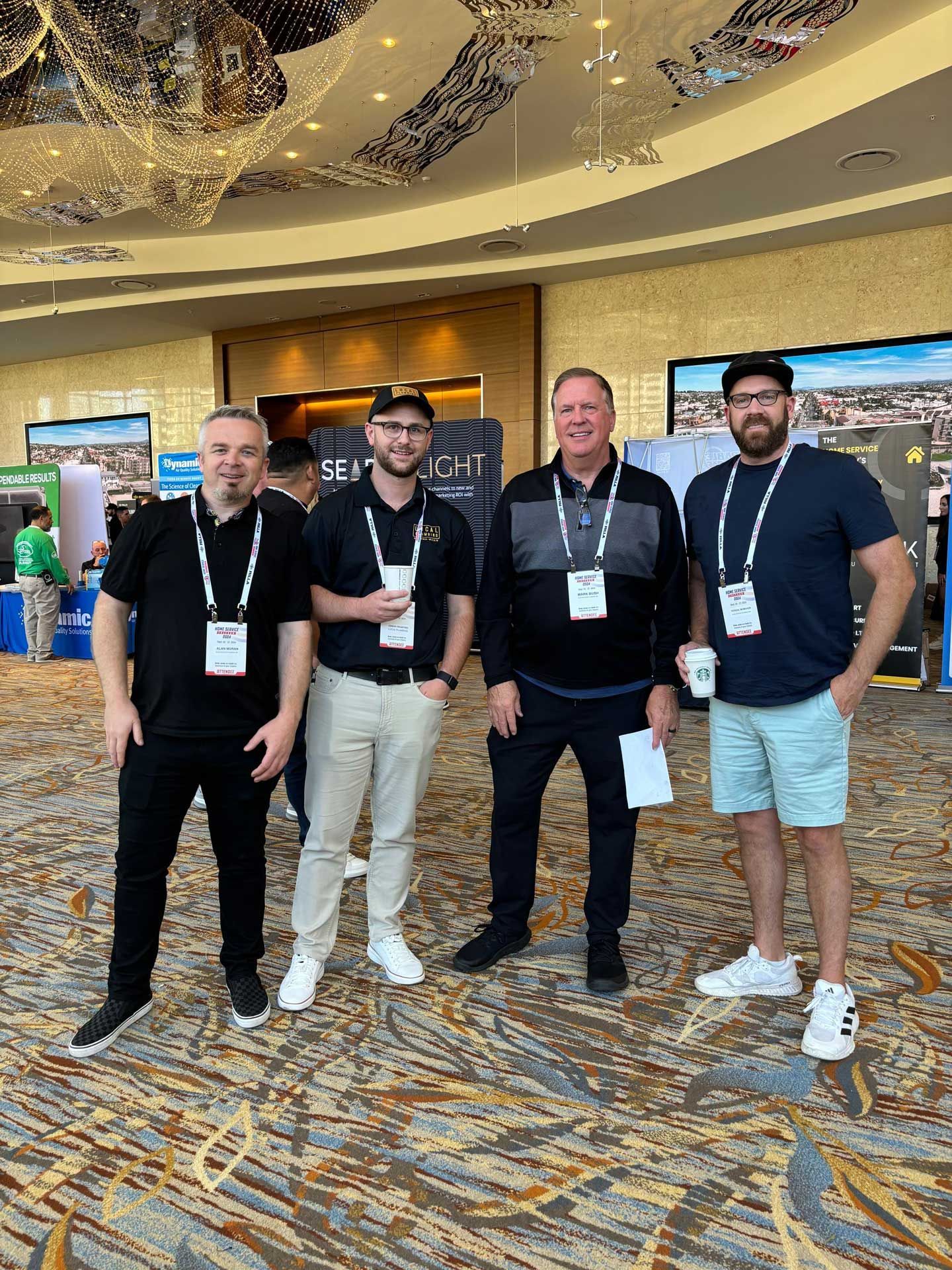Four men with lanyards pose at an event. They are standing indoors with a patterned floor.