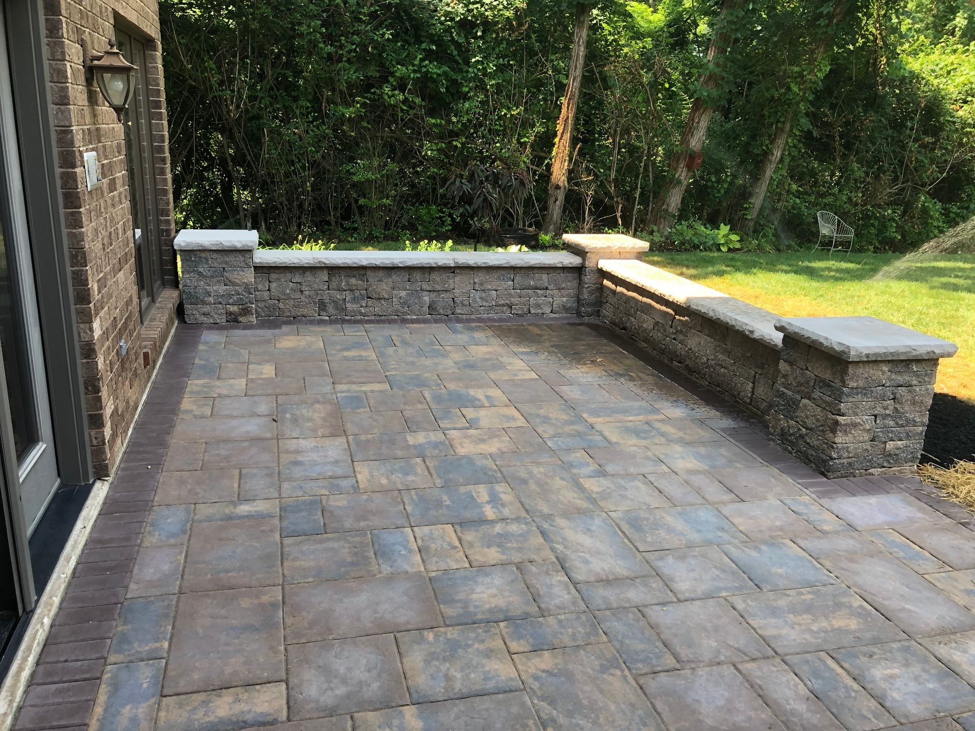 Patio with brick pavers and low stone wall, beside house with green yard.