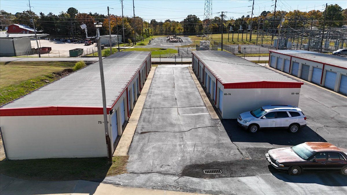 Storage units with red trim, parked cars, asphalt drive, and utility poles in an outdoor setting.