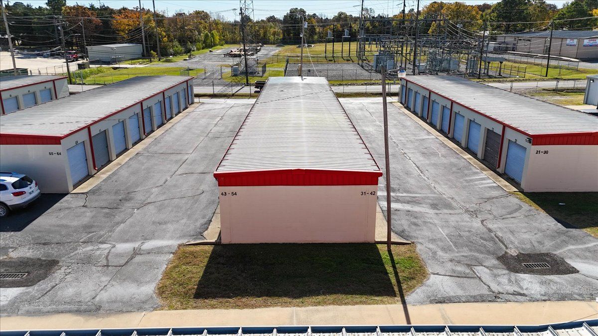 Storage units with red trim, parked cars, and power lines under a blue sky.